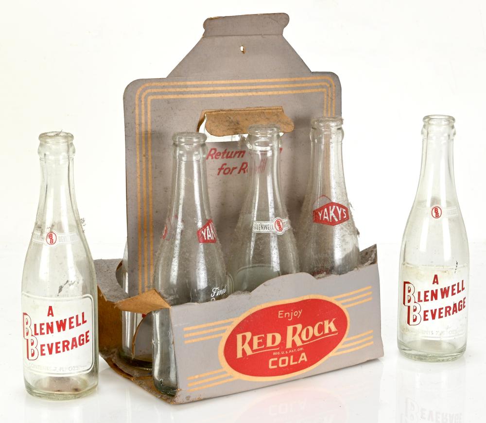 Vintage red and beige cardboard six-pack bottle carrier with four glass cola bottles on a white background.