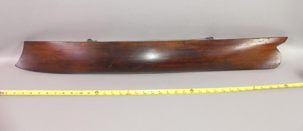 Wooden half-hull ship model section on a white background, shown lengthwise with a yellow measuring ruler beneath it.