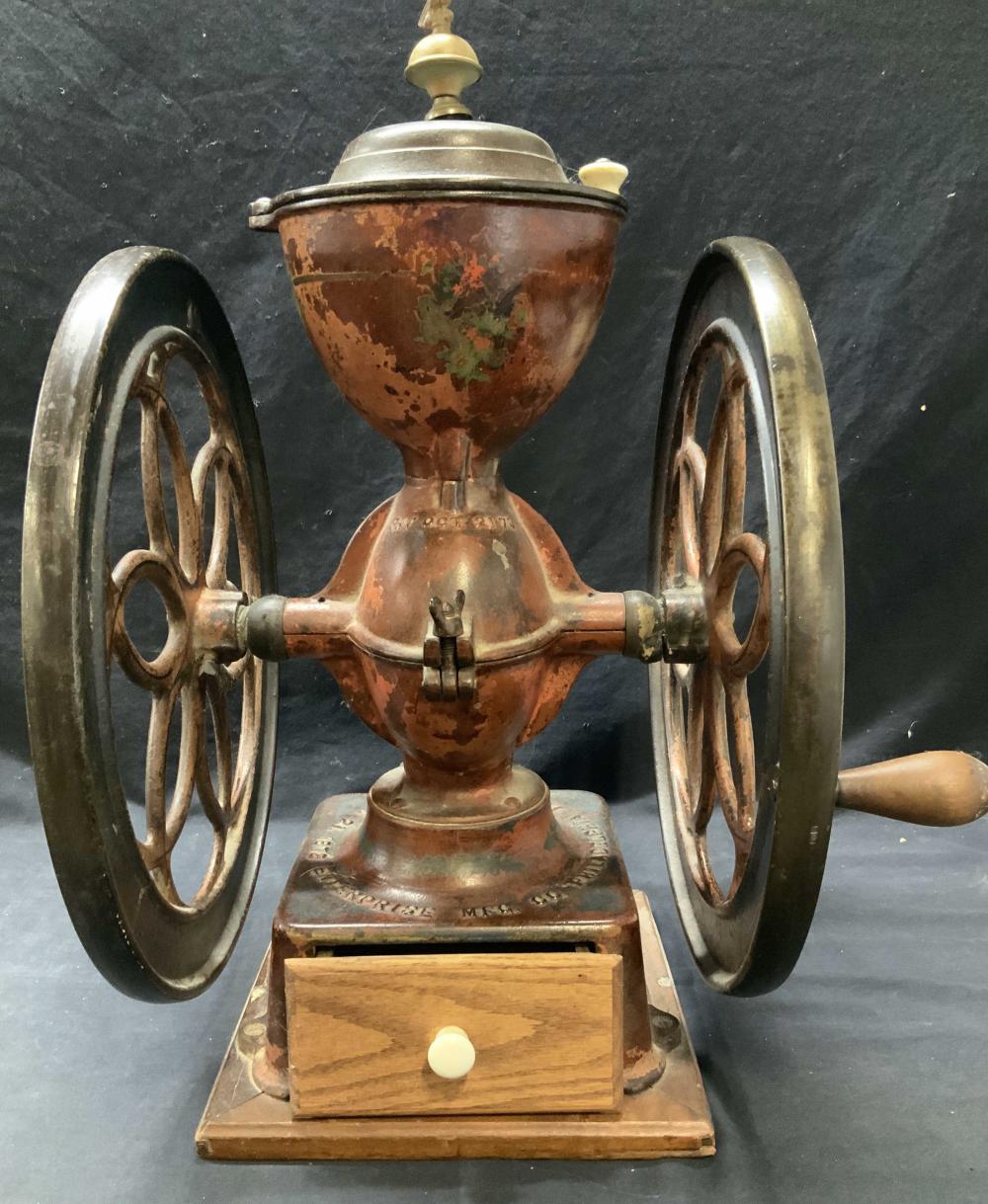 Antique-style hand-crank coffee grinder with copper-colored hopper and two spoked flywheels on a wooden base, photographed against a dark background.
