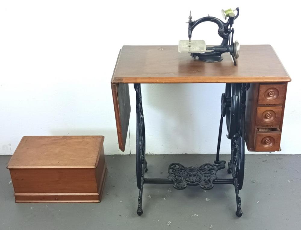 Antique black sewing machine mounted on a wooden table with drawers, shown with a wooden box beside it.