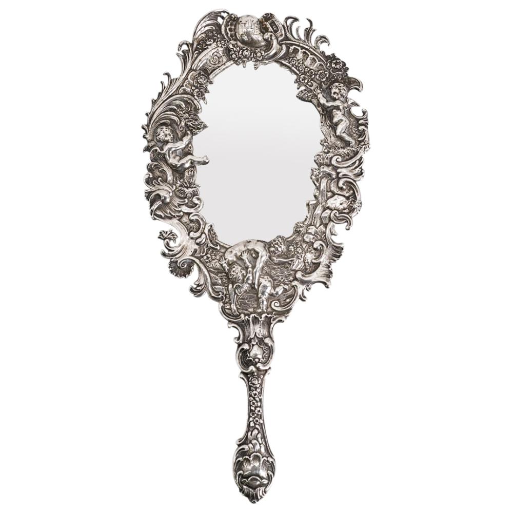 Victorian-style silver hand mirror with an oval frame and ornate cherub reliefs around the border and handle.