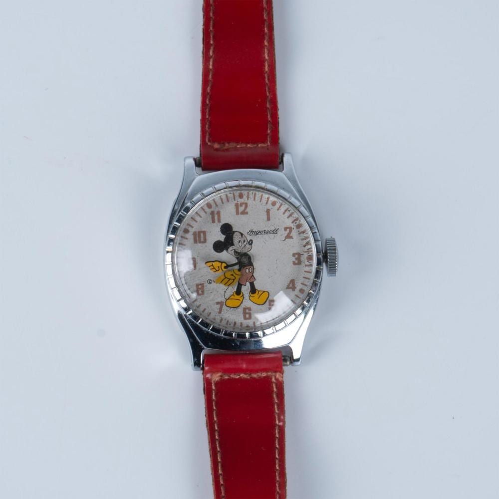 Vintage wristwatch with a red leather strap and a round silver case. The white dial shows Mickey Mouse in black ears and yellow pants with numbers around the is