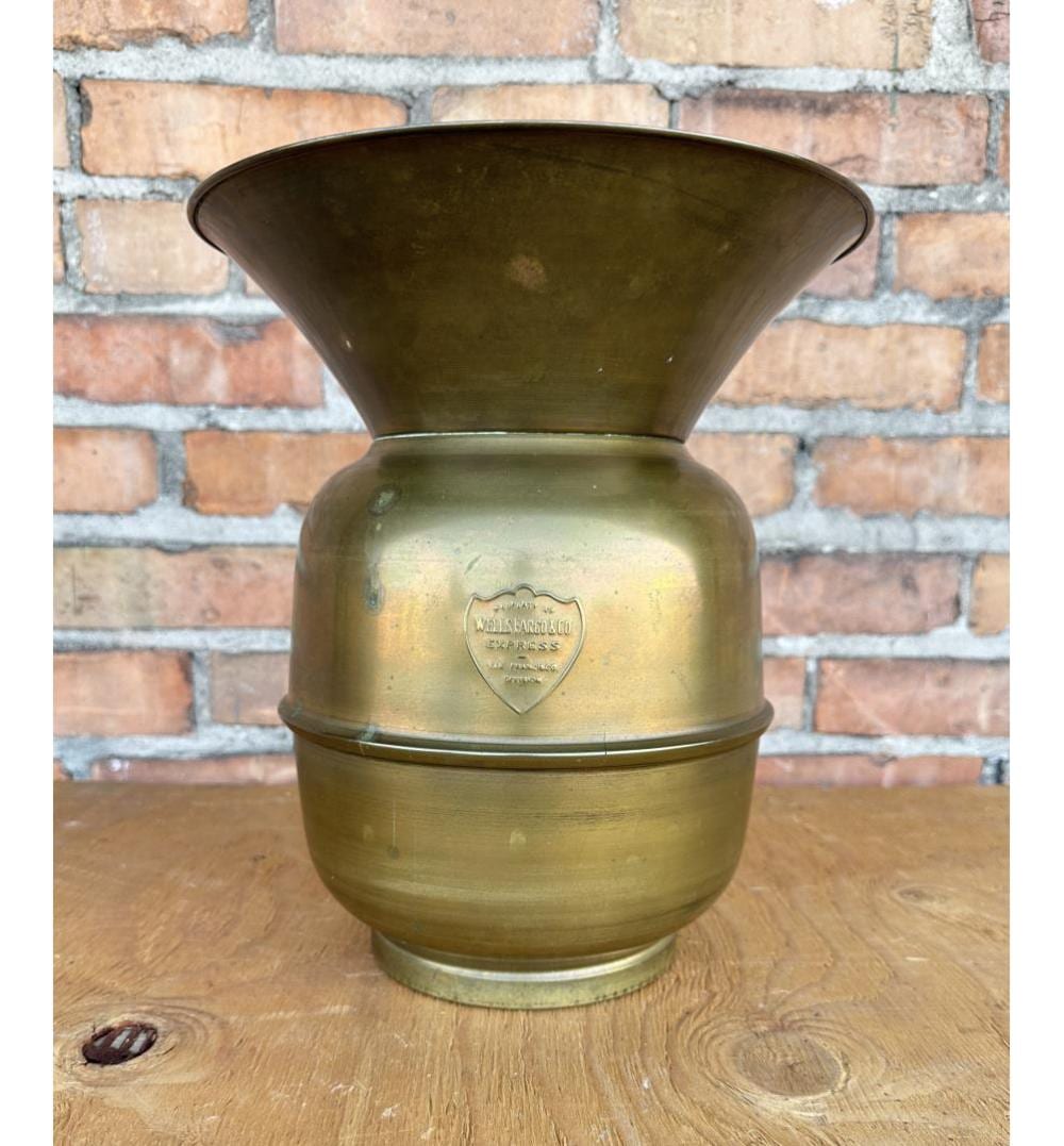 Brass spittoon with a flared funnel top and a shield-shaped logo on the front, shown against a brick wall background.