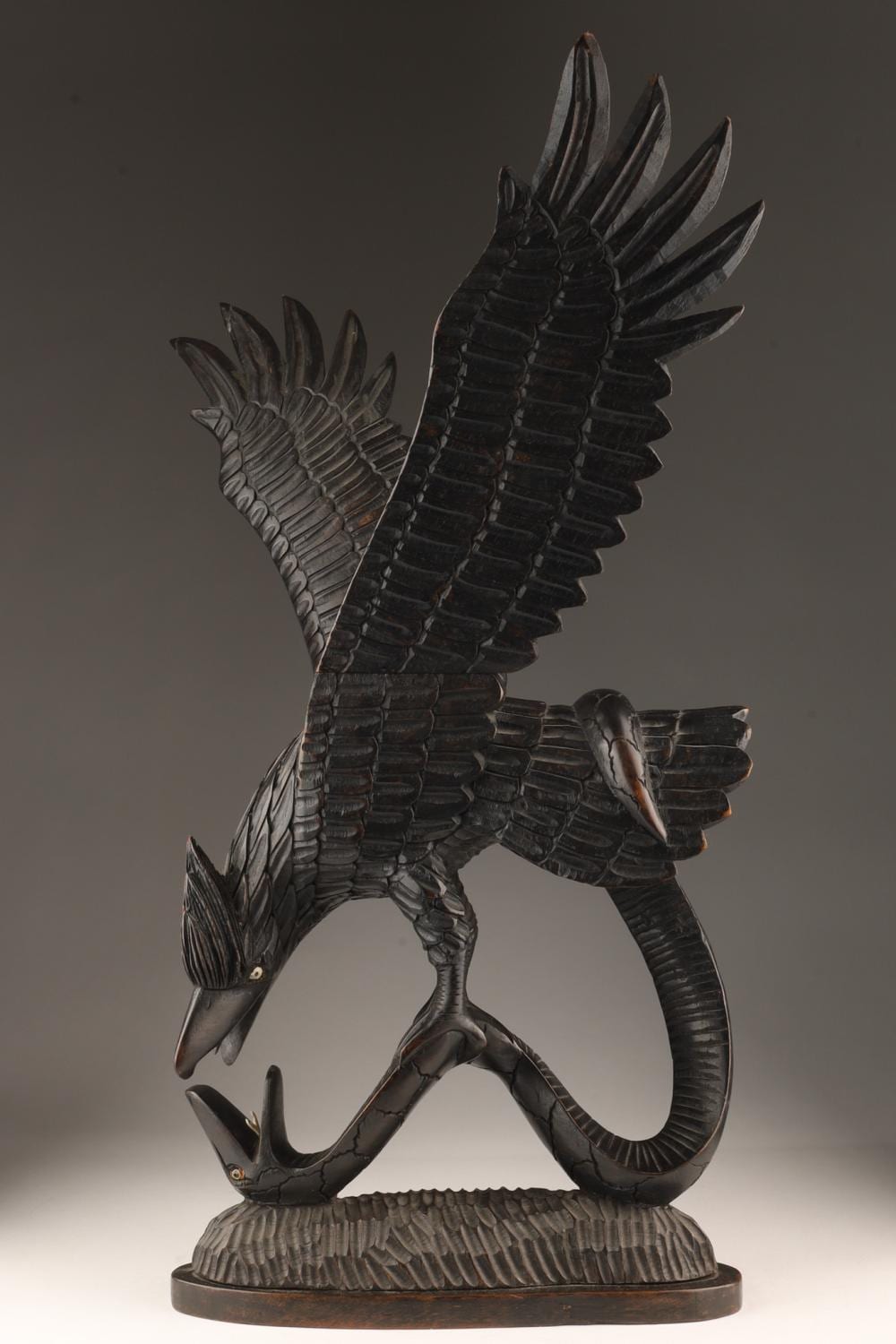 Folk art style carved sculpture of a dark bird with outstretched wings and a curved snake-like body on a base.