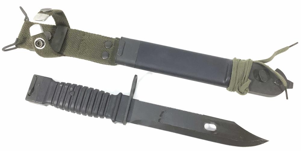 Dark metal bayonet with a serrated grip section, wrapped in a cloth strap and attached to a black scabbard.