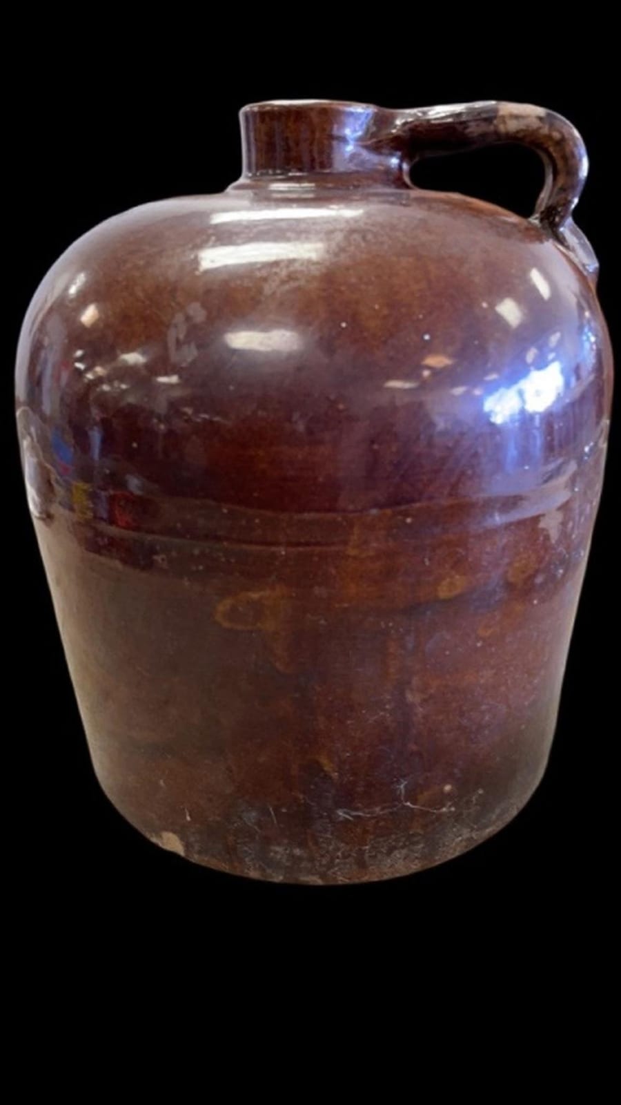 Brown glazed ceramic whiskey jug with molded body and arched handle on a dark background.