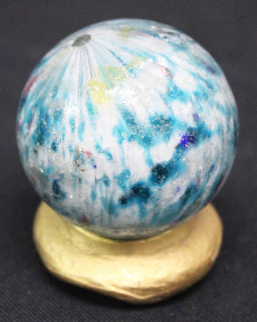 Close-up of a transparent blue and white glass marble with swirled colors resting on a small base.