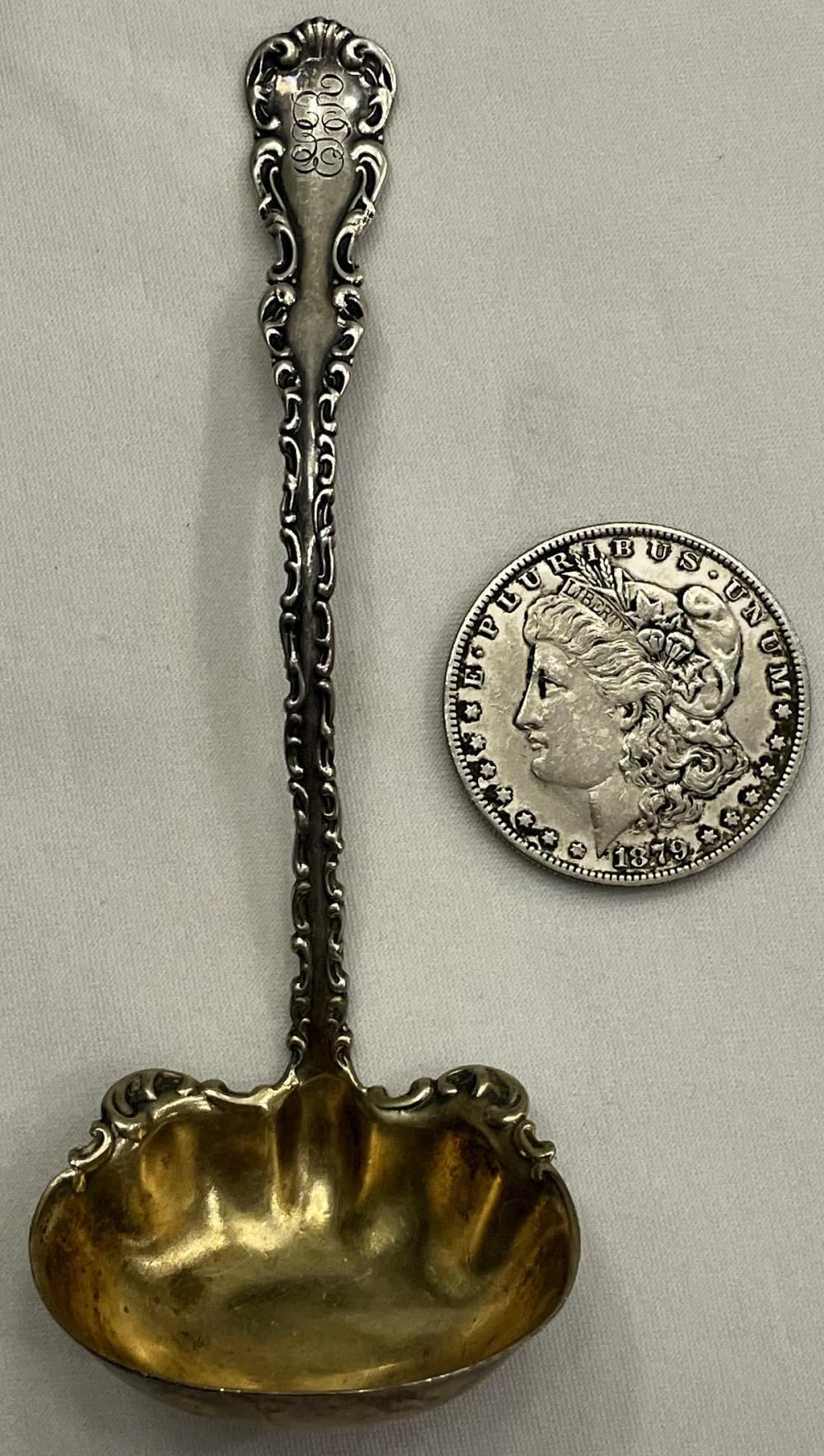 Close-up of an antique ornate oyster ladle with a silver-toned stem and gold-toned oval bowl, shown with a U.S. coin for scale.