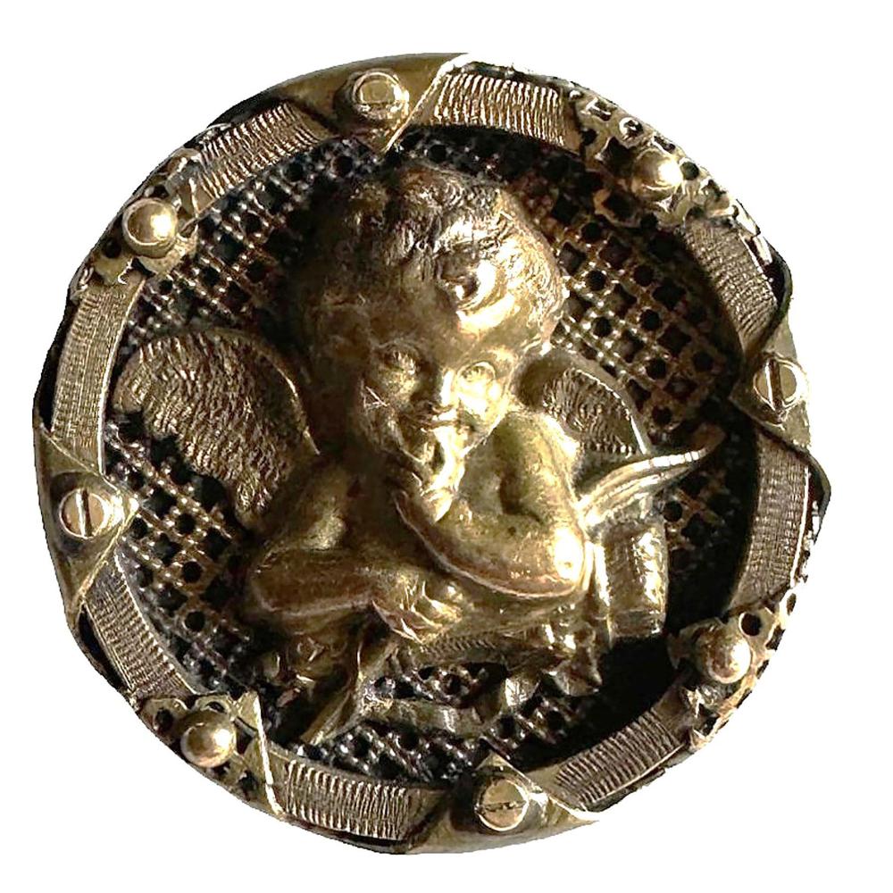 Brass round pin with a relief of a cherub (cupid) face and curly hair, surrounded by decorative geometric trim.
