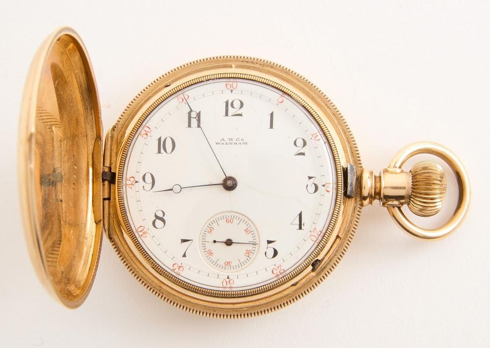 Open gold-tone pocket watch showing a white dial with Arabic numerals, black hands, and a small seconds subdial; case and crown visible.