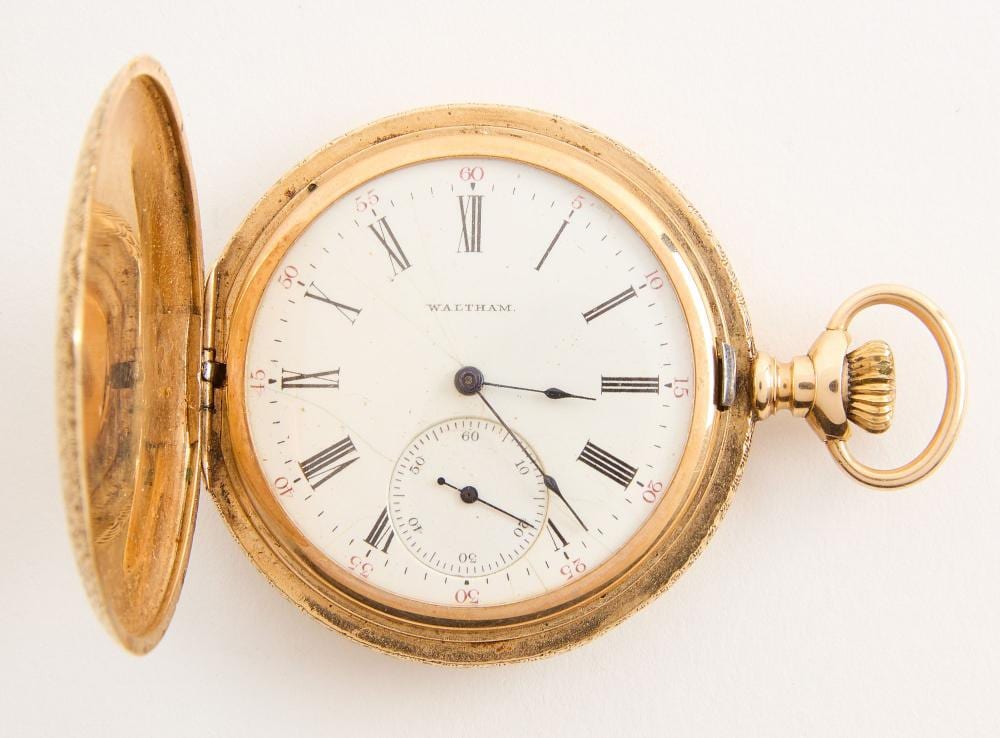 Gold-colored open-face pocket watch with roman numerals and a Waltham dial, showing a separate cover and key-wind style crown.