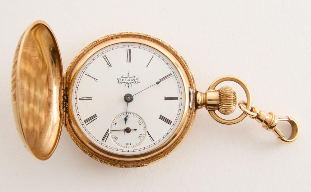 Open gold pocket watch with a white dial, Roman numerals, and a small seconds dial, shown with its key-wind stem and chain attachment.