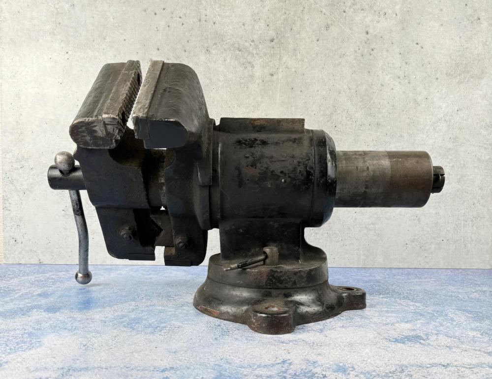 Large black bench vise with a threaded screw and metal jaw clamp mounted on a base, photographed against a light wall.