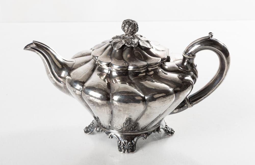 Silver teapot with a detailed lid finial, curved spout, and ornate base on a white background.