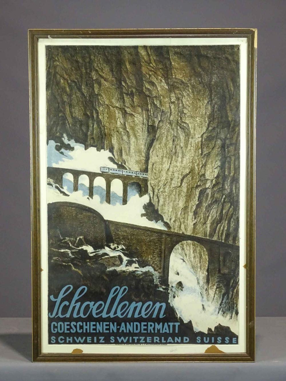 Framed Swiss travel poster showing a mountain gorge with a bridge and a train, with the word “Schöllenen” and “Schweiz/Switzerland.”