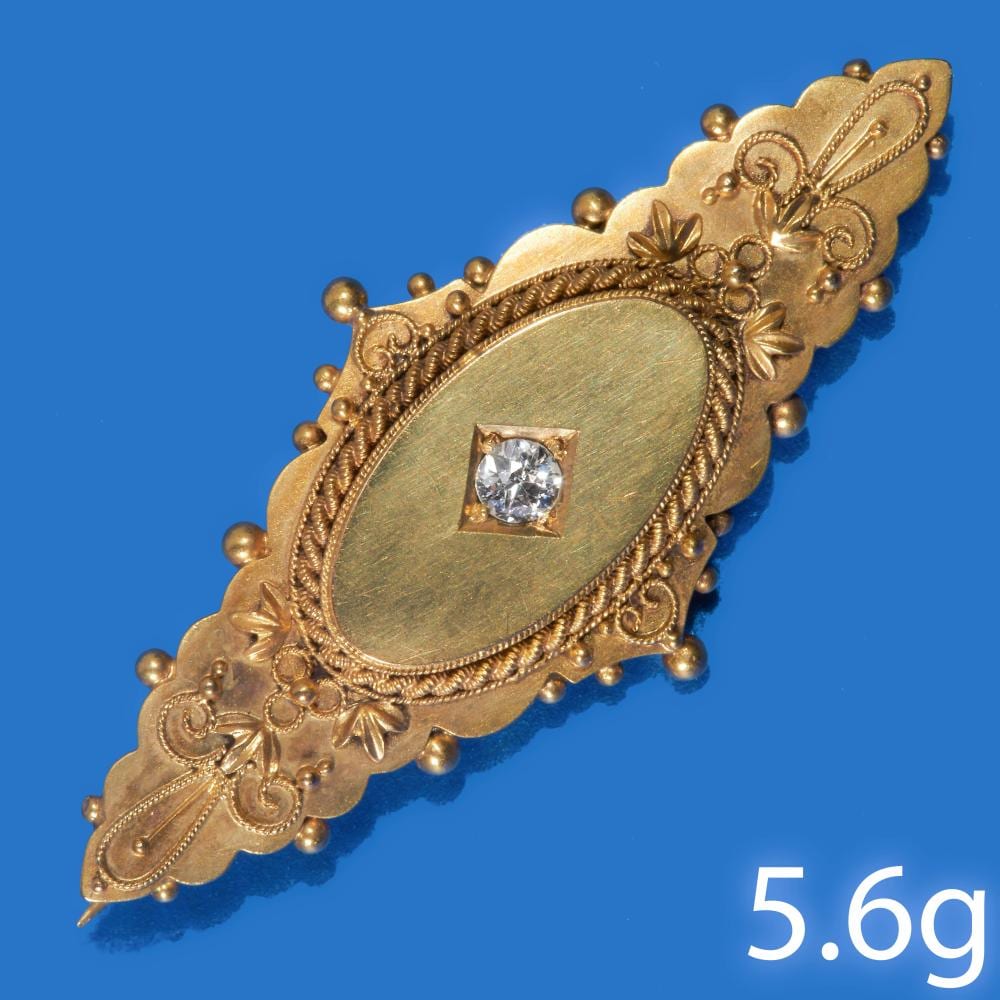 Gold Victorian-style oval brooch with ornate scrolling details and a square-set diamond in the center against a blue background.