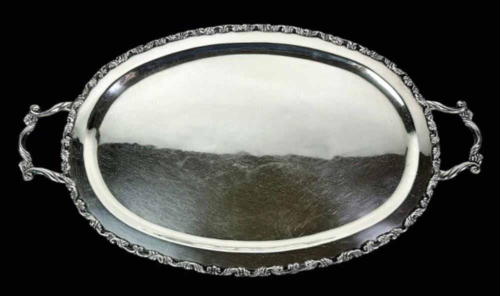 Close-up of a large silver serving tray with ornate embossed border and two curved side handles, photographed on a dark background.