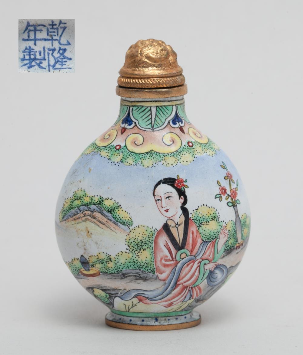 Decorative Chinese snuff bottle with a gold metal cap and pastel painted scene of a woman holding a flower by trees and birds.