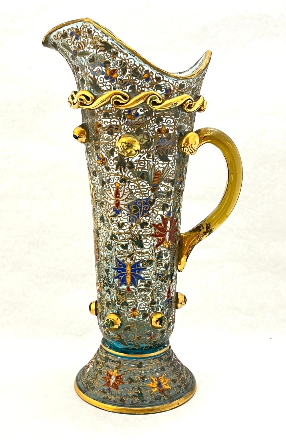 Tall enameled metal pitcher with ornate gold filigree and floral scrollwork in blue, red, and white, with curved spout and handle.