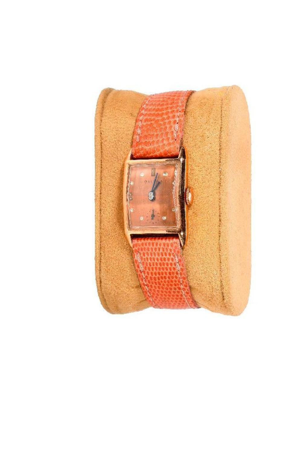 Vintage wristwatch with a square brown dial and red leather strap, shown in a front view on a tan display cushion.