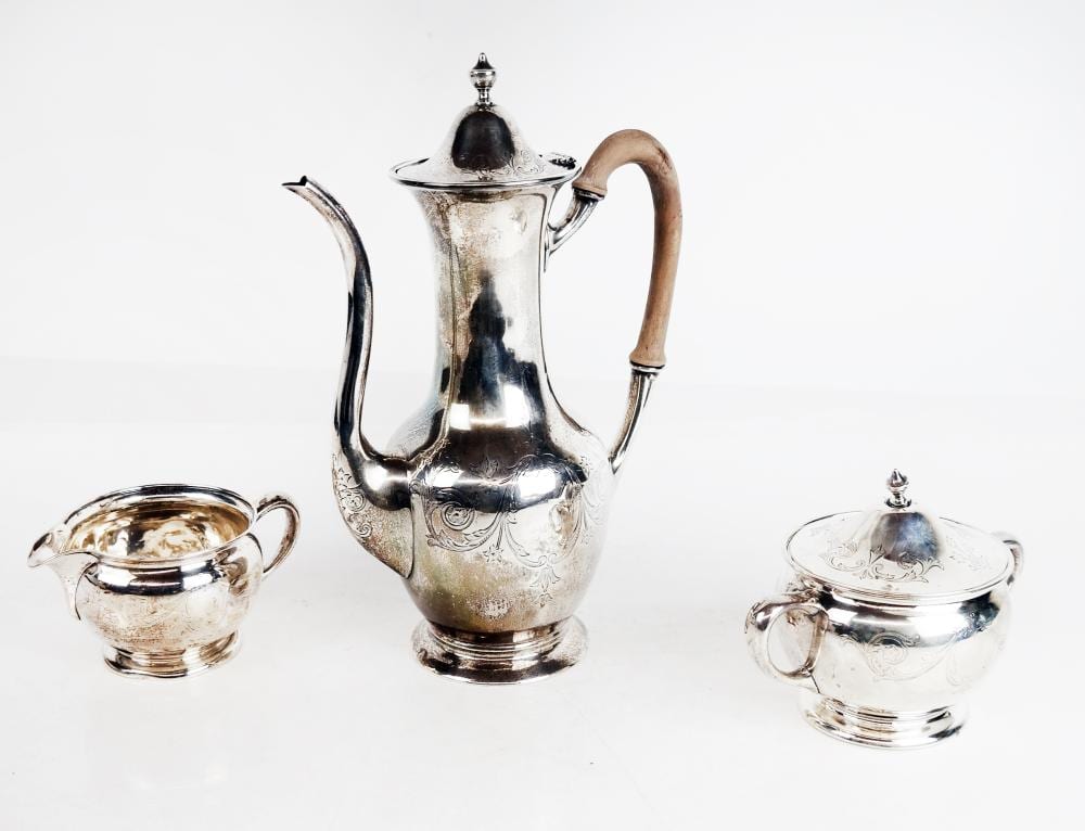 Three-piece silver tone tea set with a lidded teapot, covered creamer pitcher, and covered sugar bowl with ornate engraving and finials.