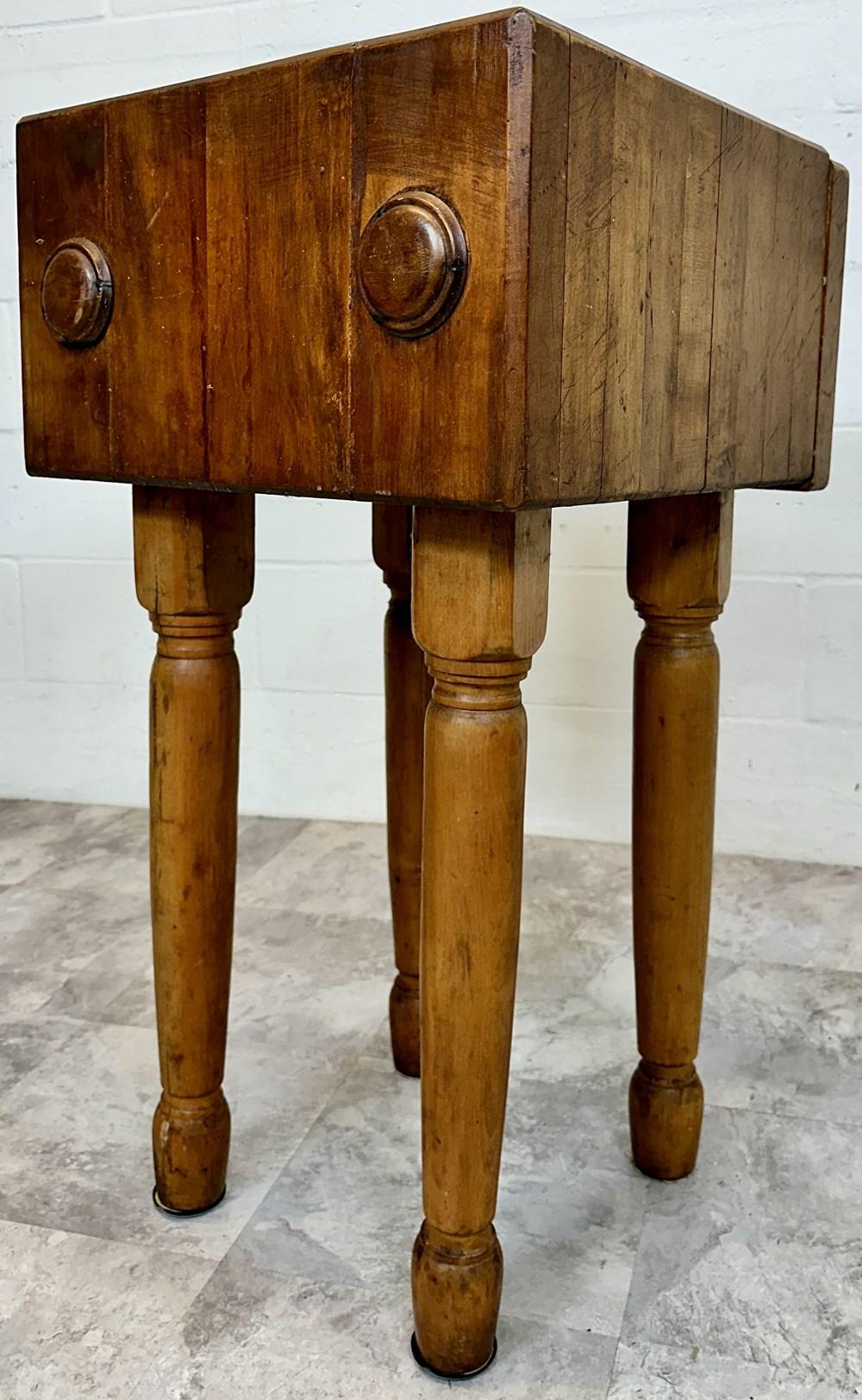 Antique wooden butcher block table with a heavy top and three turned legs, featuring metal ring handles on the apron.
