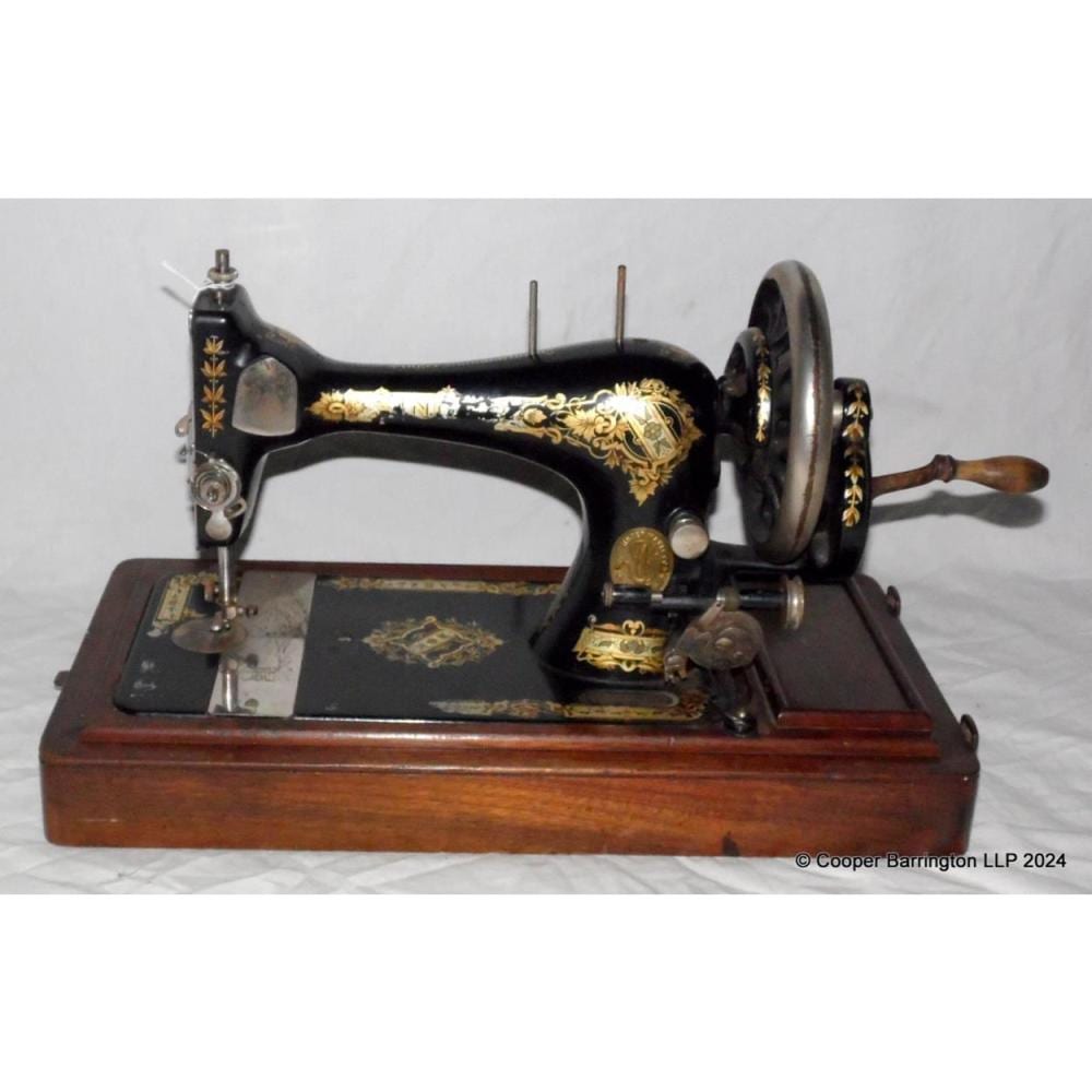Victorian-style black sewing machine with gold decorative scrollwork on a wooden base, viewed from the side with the hand wheel and needle bar visible.