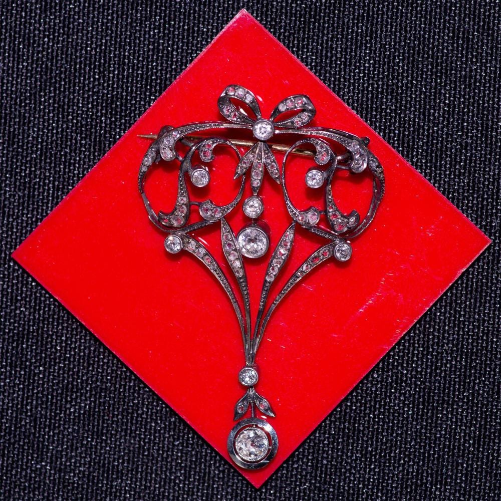 Silver-toned brooch with an oval bow and swirling vine design set with small round stones on a red square background.