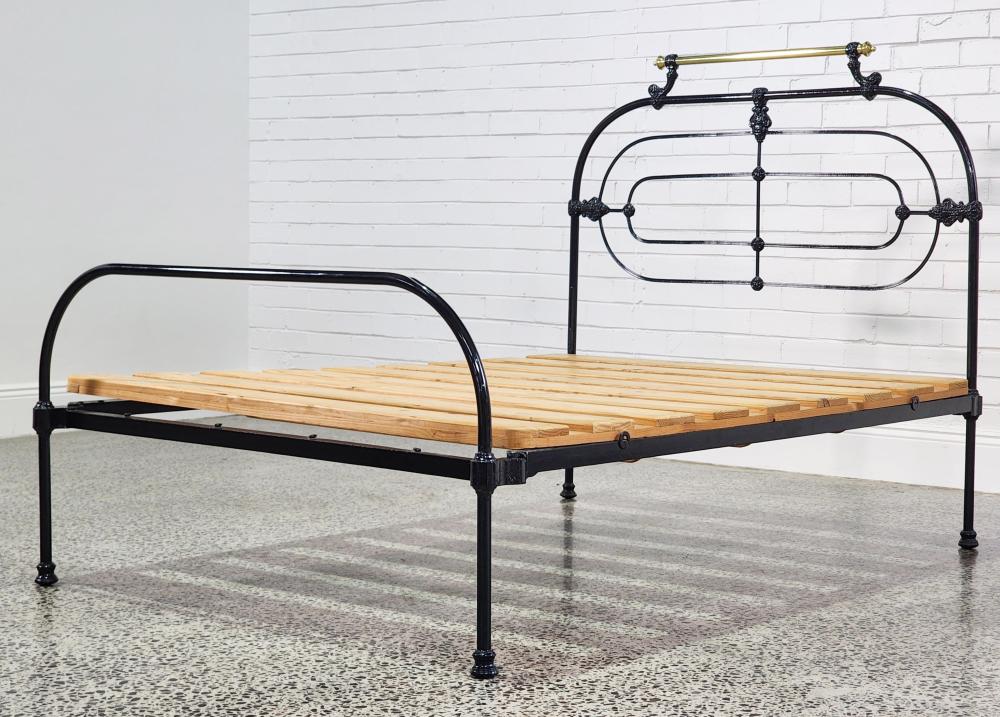 Black metal bed frame with brass-colored finials and a wooden slat platform against a white brick wall.