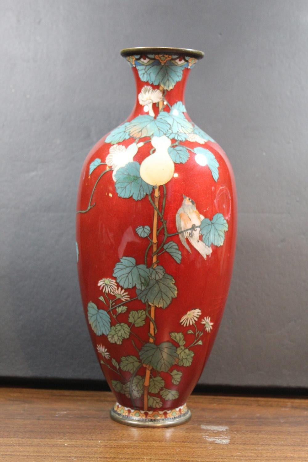 Red enamel cloisonné vase with blue leaves, white flowers, and two birds, shown front view on a wooden surface.