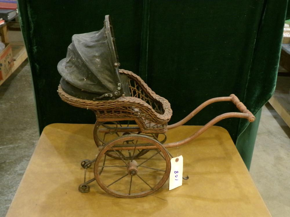 Antique-style doll buggy with a woven basket seat and visible wire wheels on a yellow display surface.