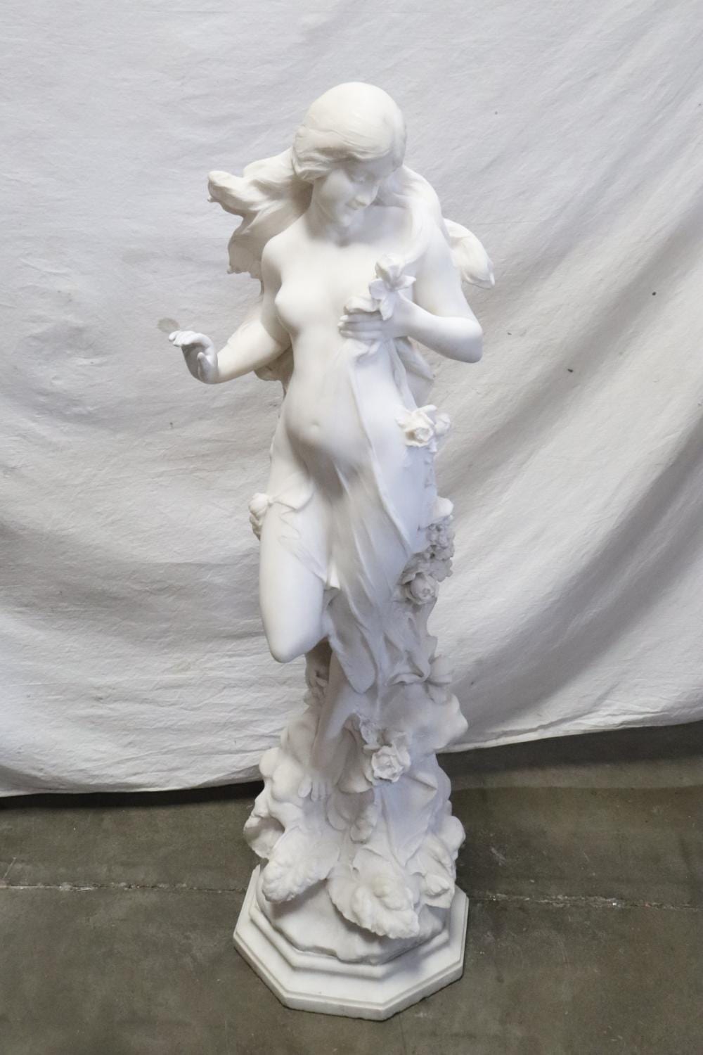 White stone sculpture of a winged young goddess holding a bouquet, with flowers carved around her pedestal.