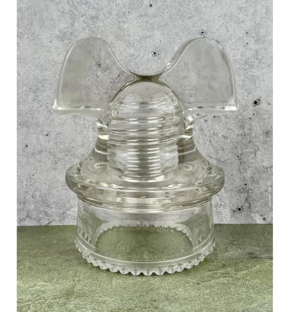 Clear glass insulator with a mushroom-like top and fluted base, shown on a green surface against a gray background.
