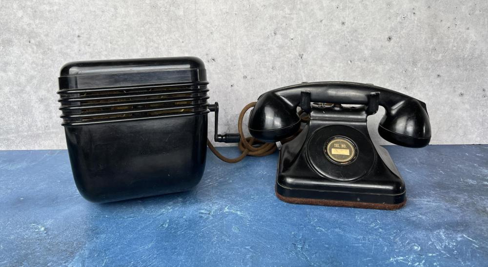 Black rotary crank telephone base with cord, shown next to its matching handset on a blue surface.