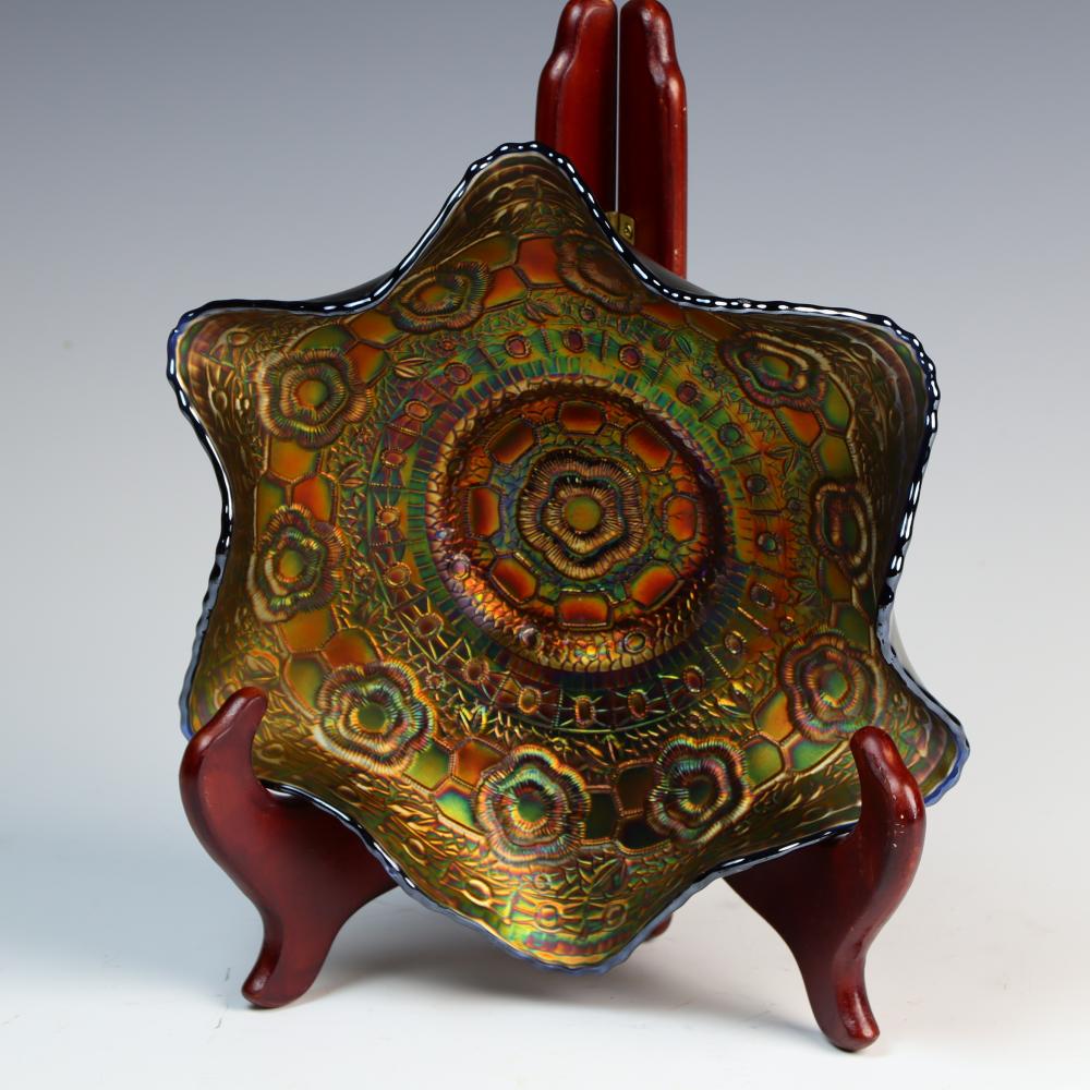 Carved carnival glass bowl in amber and green with ornate fan-shaped pattern and blue rim, shown on red stand legs.