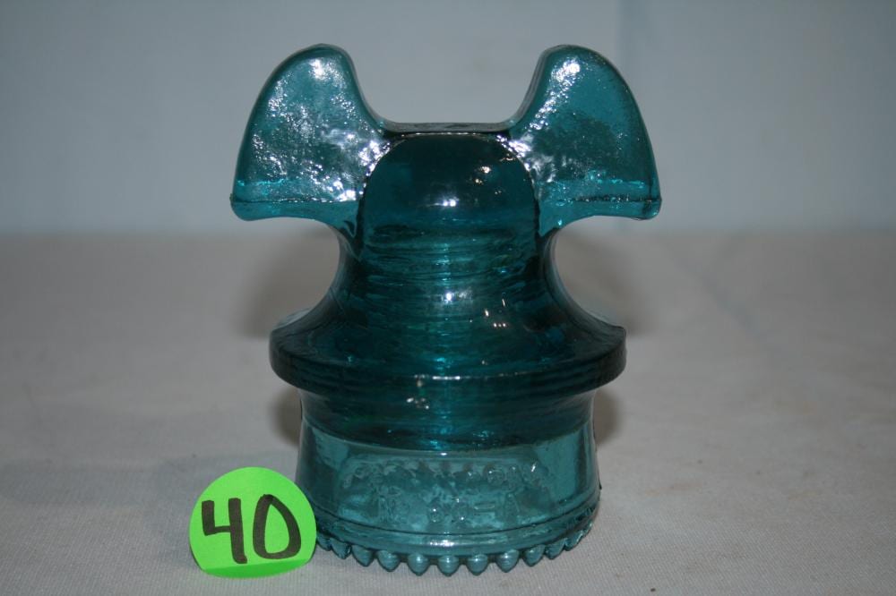 Aqua blue glass insulator with a Mickey Mouse–shaped top profile and small bead-like ring near the base on a light background.