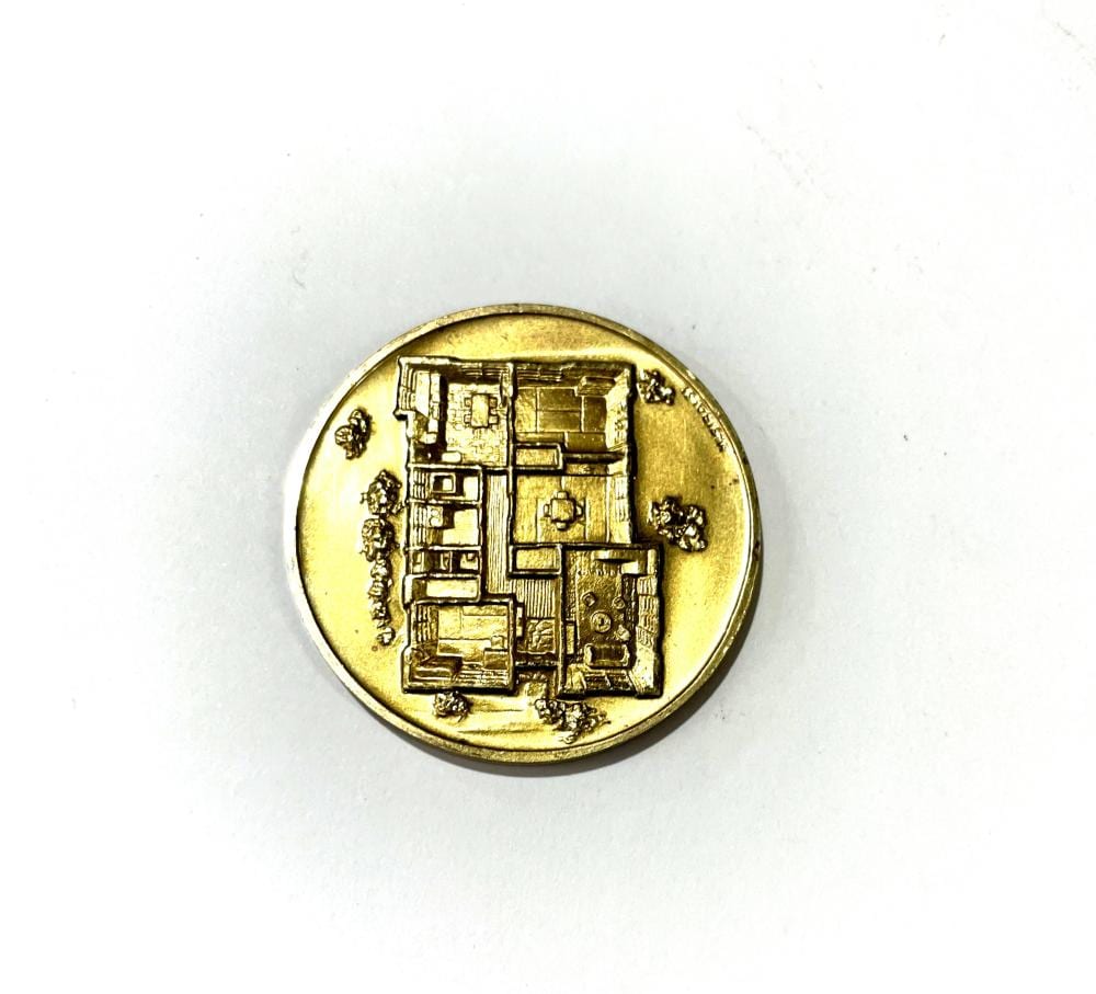 Gold-colored commemorative coin with an embossed architectural design and small figures along the sides, shown on a white background.