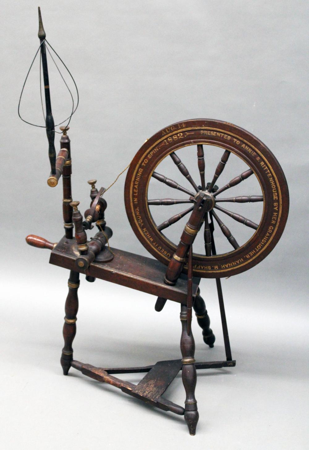 Antique wooden yarn spinning wheel with circular wheel, multiple wooden drive posts, and a hanging fiber bowl on a stand against a light background.
