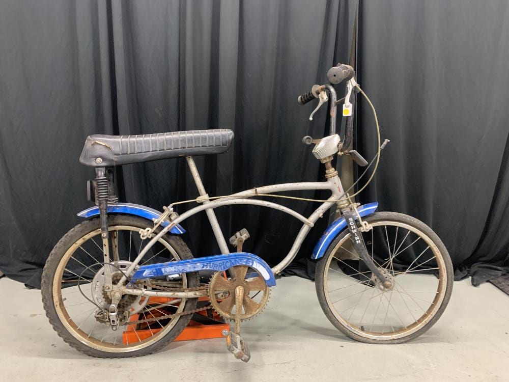 Vintage white BMX bicycle with blue fenders and a blue frame trim, shown side-on against a dark curtain backdrop.