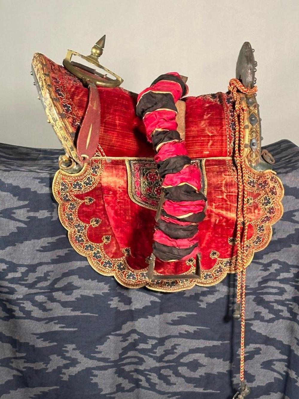 Red camel saddle with ornate gold and floral embroidery, side panels, and a strap and reins on a blue patterned surface.