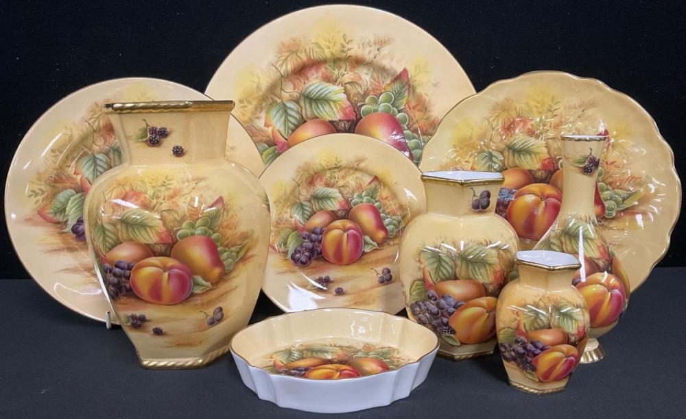 Collection of decorative porcelain fruit-pattern vases, bowls, and plates with gold handles and trim against a dark background.