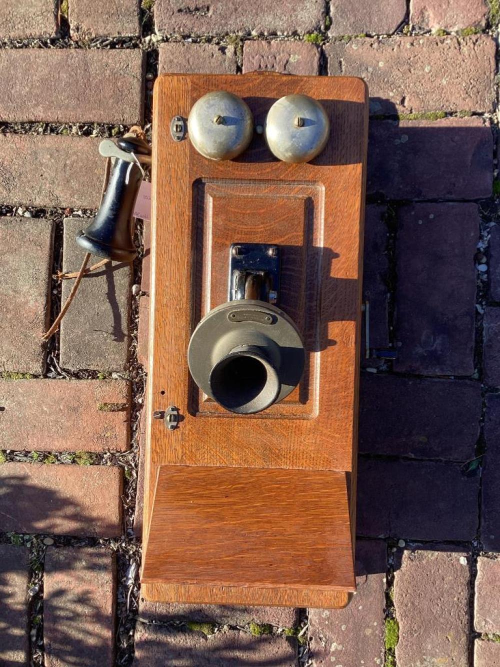 Wooden wall-mounted telephone case with two metal knobs at the top and a circular handset holder in the center, photographed outdoors against stone steps.
