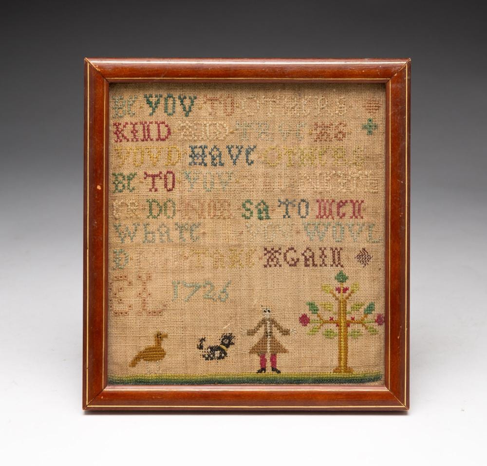 Framed needlepoint sampler with alphabet and a small figure by a flowering tree, stitched on tan fabric.