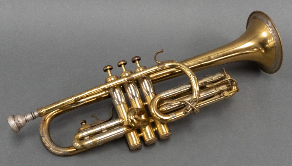 Brass trumpet with multiple valves, coiled tubing, and a flared bell, photographed against a gray background.