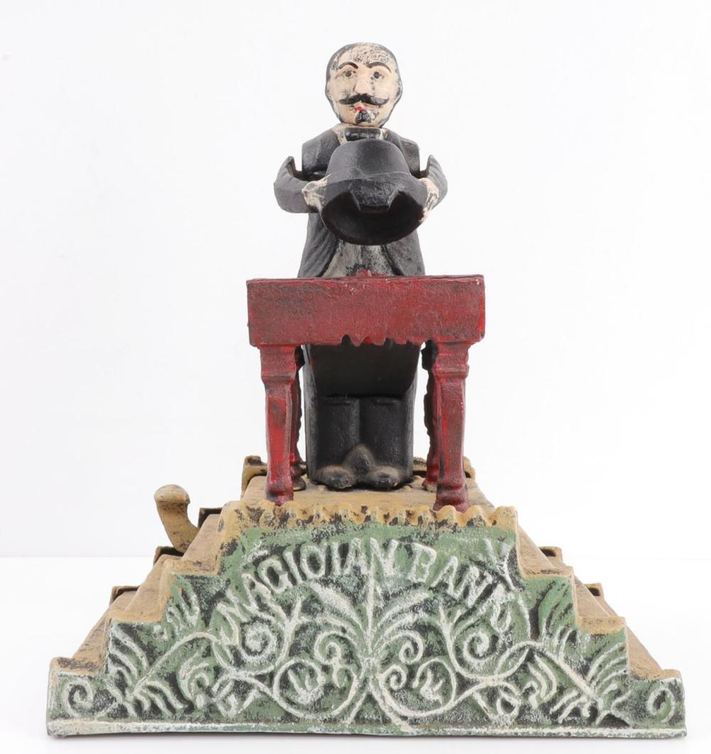 Cast iron mechanical magician-style piggy bank figure holding a coin bucket on a decorated base, photographed against a white background.