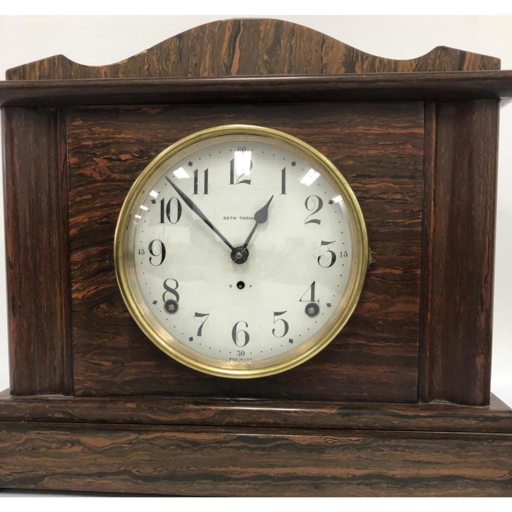 Close-up of an antique wooden mantel clock with a round white dial and gold-toned bezel, showing black roman numerals and hands.
