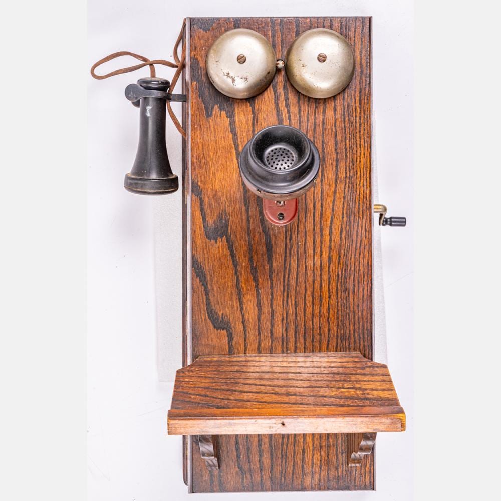 Antique wall-mounted telephone with two silver bells and a black handset cradle on a carved wooden board.