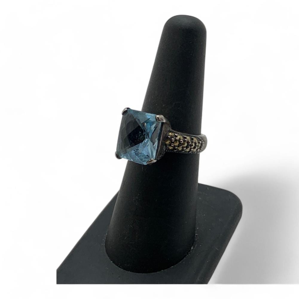 Sterling silver ring with a square blue gemstone set in a prong setting, shown on a black display stand.
