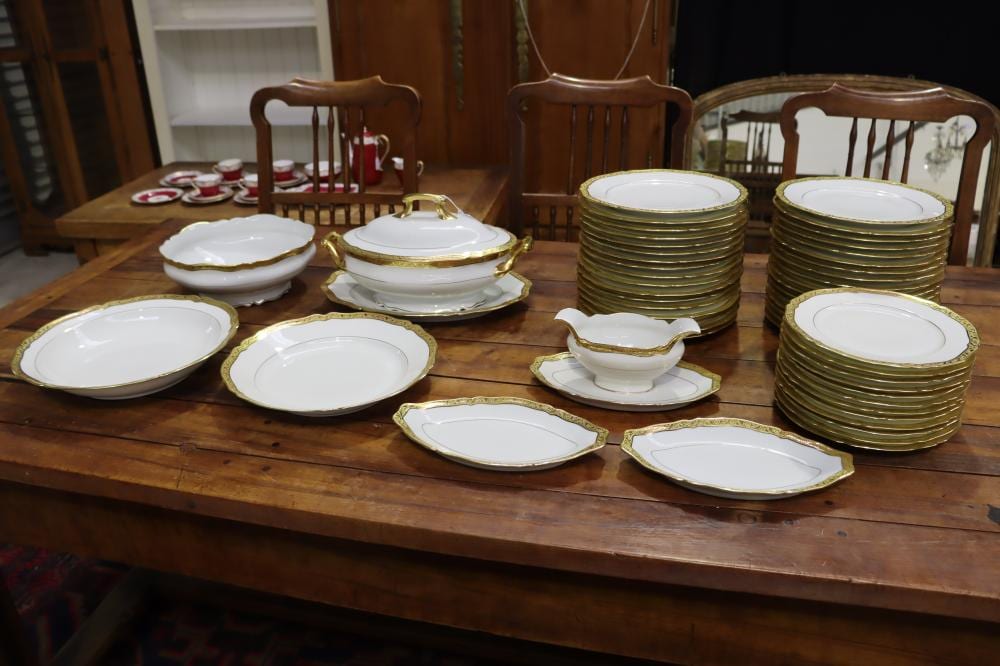 Stacked white porcelain dinner plates and serving dishes with gold trim on a wooden table in a room.