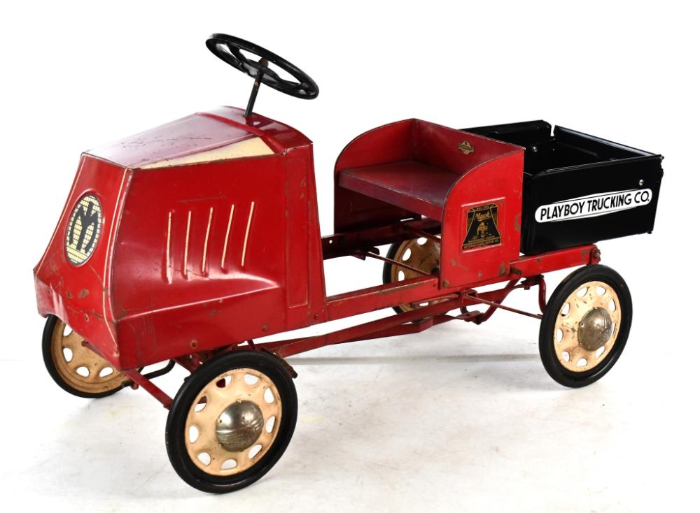 Red pedal car styled like a dump truck with black seat, steering wheel, and a black cargo bed labeled “PLAYBOY TRUCKING CO.”